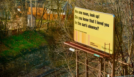 A billboard on a sunny winter day, with leafless trees and some vegetation. Train tracks intersect the photograph. The billboard, mostly in yellow, features a haiku, credited to Jason Livingston and Phoebe A. Cohen: “No one noon, look up! Do you know that I loved you in the sun’s absence?” Vertical lines in gradients of orange and yellow are next to the hashtag #InthePathofTotality, and Squeaky Wheel’s logo. 