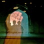 Still image from Kalpana Subramanian's film "Incantation" (2021). A photograph depicting multiple images projected on a surface. The shadows of what look like two young people, arms on each other’s shoulders, standing under an arched gateway to a historic building in Delhi, India. Upon this image is an extended hand, palm open. It looks like the young people are in the palm. 