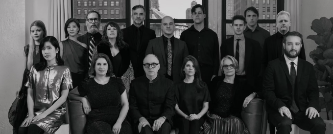 The 15 members of the Talea Ensemble pose together for a black-and-white group portrait. 