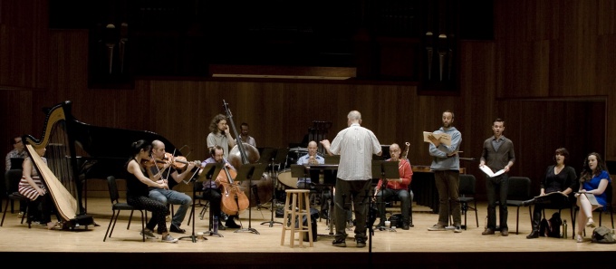 A contemporary music ensemble rehearses onstage in a concert hall, with a conductor standing at center facing seated instrumentalists&mdash;strings, harp, winds, and percussion&mdash;while vocalists stand to the right holding scores during a run-through. 