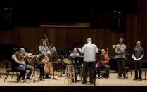 A contemporary music ensemble rehearses onstage in a concert hall, with a conductor standing at center facing seated instrumentalists&mdash;strings, harp, winds, and percussion&mdash;while vocalists stand to the right holding scores during a run-through. 