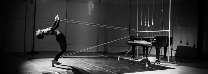 artistic photo of an avant-garde performance where a dancer an in evocative pose has long strings tied to a suspended piano. 