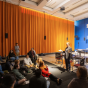 A group of students and community members sit in a semicircle on the floor and in chairs of a large room with tall orange curtains. A speaker stands at the front, gesturing while addressing the group. A piano, tables, and a large projection screen are visible in the background. 
