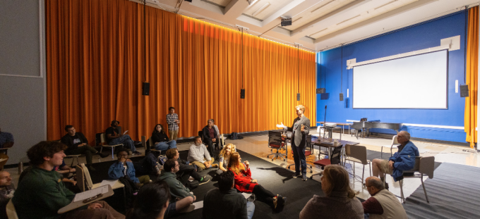 A group of students and community members sit in a semicircle on the floor and in chairs of the Experimental Intermedia Space in Slee Hall with tall orange curtains. A speaker stands at the front, gesturing while addressing the group. A piano, tables, and a large projection screen are visible in the background.
