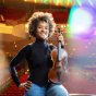 Melissa White stands on a concert hall stage holding a violin, smiling toward the camera. Colorful stage lights create lens flares around her, and rows of red theater seats are visible in the background. 