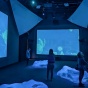Visitors explore an immersive multimedia art exhibit in a darkened room illuminated by large blue projection screens displaying abstract, floating organic forms. Sculptural, mound-like structures on the floor emit soft light, and people move around or observe the installation. The scene is part of the exhibit “Serenades for Settling: Tending Ostreidae” at the Burchfield Penney Art Center. 