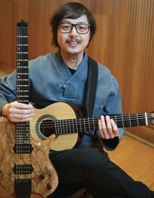Sungmin Shin is holding two guitars. In his left hand, he holds the neck of an electric guitar with a natural wood-grain body and visible pickups. Across his lap, he holds an acoustic guitar with a light wood finish, resting his right hand near the strings and his left hand on the fretboard. A black guitar strap runs over his shoulder. He faces the camera directly with a relaxed, friendly expression. 