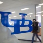 The UB logo displayed on a transparent glass wall with a student walking by in the background. 