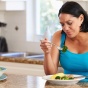 A frustrated or disappointed-looking woman eating a healthy meal including broccoli in a kitchen setting. 