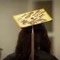 An ALANA graduate shows their decorated cap with the phrase "WE RISE TO LIFT OTHERS.". 