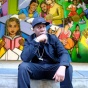 Grandmaster Flash sits on a curb with a colorful mural in the background. 