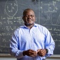 Herbert Foso stands in front of a chalkboard covered with physics equations. 
