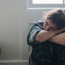 A sad looking young person holds a cell phone while resting their head on their folded arms. 
