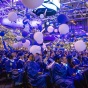 Students at a 2023 College commencement story sit, gazing up in awe at balloons dropping down from the ceiling overhead. 