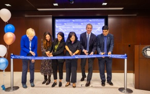 six people standing next to each other cutting blue UB ribbon. 