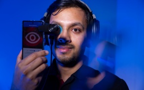 headshot of man with headphones holding phone up to right eye with blue background. 