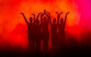 silhouette of five dancers with arms up overhead with foggy orange background. 