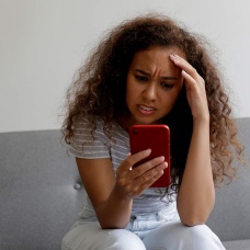 A woman sitting on a couch looking at a smart phone with a distressed look on her face. 