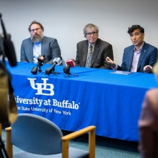 Three men sit at a table with a University at Buffalo table cloth talking with microphones and a camera. 