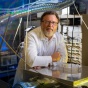 Man in white lab coat with glasses looking into camera leaning against counter in spectroscopy lab. 