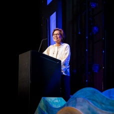 Diana Aga stands smiling at a podium on stage, wearing glasses and a light-colored blouse. She is delivering a presentation, illuminated by stage lighting. The background is dark with hints of stage equipment and blue decorative elements in the foreground. 