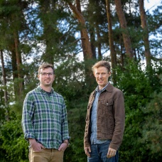 Two men standing outdoors in front of a backdrop of tall trees and greenery, smiling at the camera. Both are casually dressed, one in a plaid shirt and the other in a jacket. 