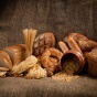 A rustic still life arrangement of various types of bread, pasta, and grains displayed on a burlap surface. The assortment includes sliced bread, loaves, baguettes, a pretzel, uncooked spaghetti, and noodles spilling from a ceramic jar. 