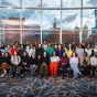 A diverse group of people seated indoors for a group photo, with a city skyline visible through large windows behind them. 