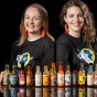 Two women wearing chicken wing earrings standing behind a row of hot sauces. 