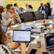 Dean Grabill, seated at the far left, participates in a roundtable discussion with conference attendees gathered around a table filled with laptops, notebooks, and meeting materials. 