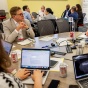 Dean Grabill, seated at the far left, participates in a roundtable discussion with conference attendees gathered around a table filled with laptops, notebooks, and meeting materials. 