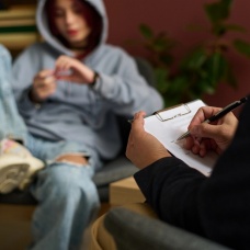 Teen sitting casually in a chair, talking with an authority figure with a paper and pen. 