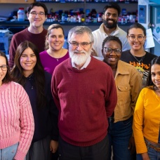 A group portrait taken inside a chemistry laboratory shows nine people standing close together and smiling at the camera. 