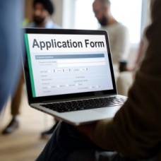 Person holding a laptop displaying an online application form while others stand in the background, representing the process of applying online. 
