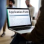 Person holding a laptop displaying an online application form while others stand in the background, representing the process of applying online. 