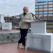 Reinhard Reitzenstein, professor of sculpture, stands outdoors at Canalside beside a granite sculpture from his exhibit &ldquo;Destination,&rdquo; with downtown buildings and rail lines in the background. 