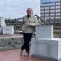 Reinhard Reitzenstein, professor of sculpture, stands outdoors at Canalside beside a granite sculpture from his exhibit &ldquo;Destination,&rdquo; with downtown buildings and rail lines in the background. 