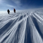 Two people walk across a vast, windswept snowy landscape, with long ridges of sculpted snow stretching toward the horizon under a deep blue sky. 