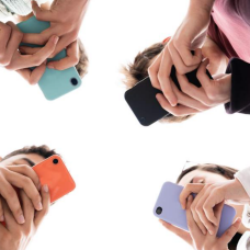 A group of students stands in a circle viewed from below, each holding a smartphone with colorful cases, their hands meeting toward the center against a bright white background. 