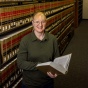 Rachael Hinkle stands in a library aisle holding an open book, surrounded by shelves of bound volumes. 