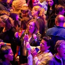 A crowd of people gathered at an indoor event under colorful purple and blue lighting, talking and reacting as they stood close together. 
