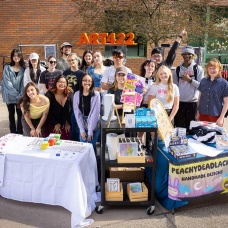 Group of students from an ART 422 class stand behind outdoor pop-up tables displaying handmade artwork and products. Signs include &ldquo;ART422&rdquo; in large orange letters behind them and a banner reading &ldquo;PEACHYDEADLACK HANDMADE DESIGNS.&rdquo; A poster on one table reads &ldquo;Student Art Sale.&rdquo; Tables are filled with prints, crafts, and small items for sale as students present their work on campus. 