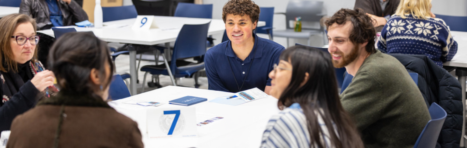 Four people sit around a small round table in a blue-walled office, engaged in discussion. A man gestures as he speaks while two women with laptops listen, and a whiteboard behind them is filled with equations, diagrams, and notes. 