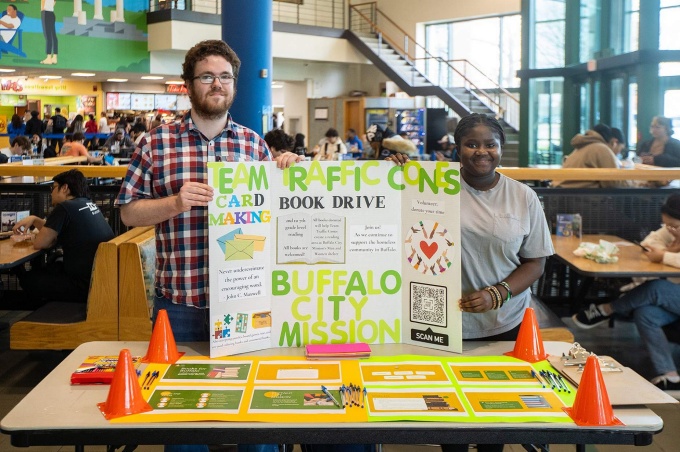 Two students stand behind a table in a busy campus commons area, holding a large poster board promoting a service project. 