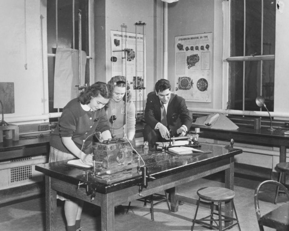 Black and white photo of Hochstetter Physics Lab in 1950. There are three people smiling and working over a lab table using equipment. 