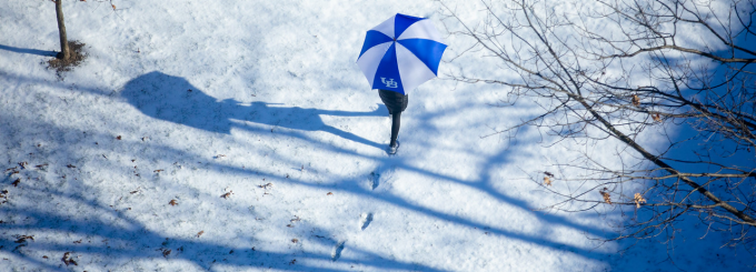Person walking in snow with a UB umbrella. 
