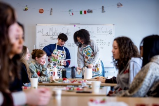 A classroom scene shows students participating in an Italian language and culture activity centered on food and drink. Several students sit around tables in the foreground and along both sides of the room, while two students stand at the front serving refreshments. 