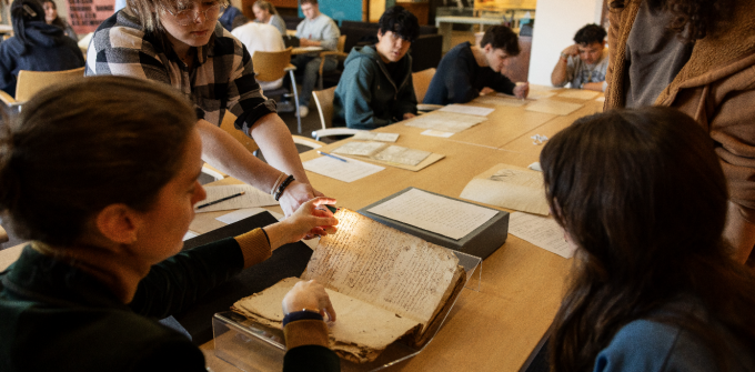 Students gather around a large table, examining rare historical documents during a hands-on archival learning session. In the foreground, several students lean over an open manuscript resting in a clear support cradle. One student carefully lifts a page while another shines a small light through the parchment or paper, illuminating the handwritten text and revealing details of the document's material and construction. 