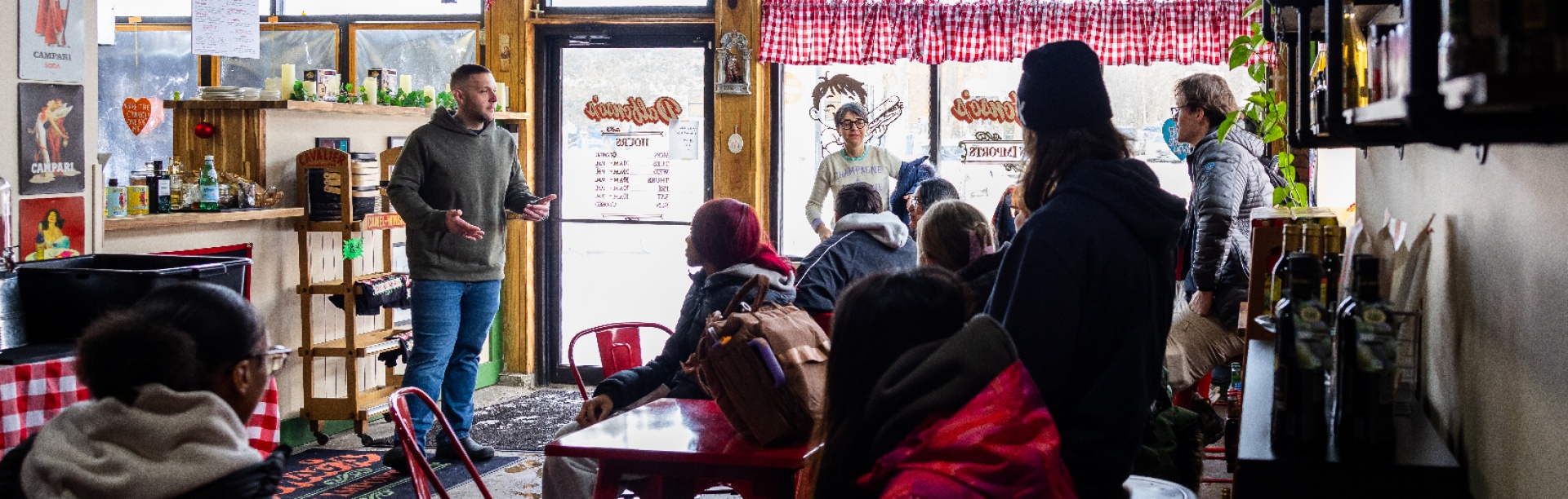 Students gather inside a small local restaurant during a class field trip, listening to a speaker near the entrance while seated at red tables. 