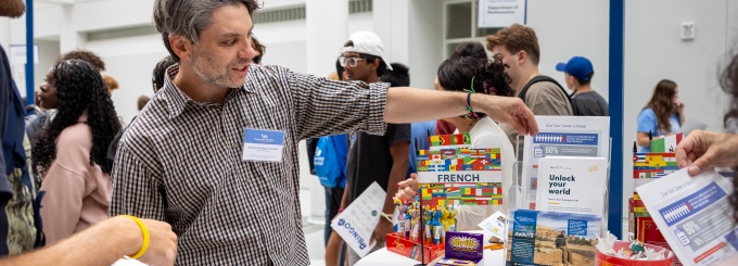 A group of prospective students gathers around a table at a university information event while a faculty member speaks with them. The setting is a bright indoor atrium with high white walls and many students in the background.